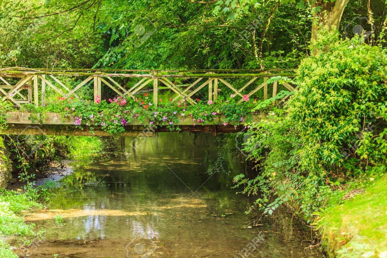 Holzbrücke mit Blumenschmuck über einem kleinen Fluss im Grünen, umgeben von üppiger Vegetation.