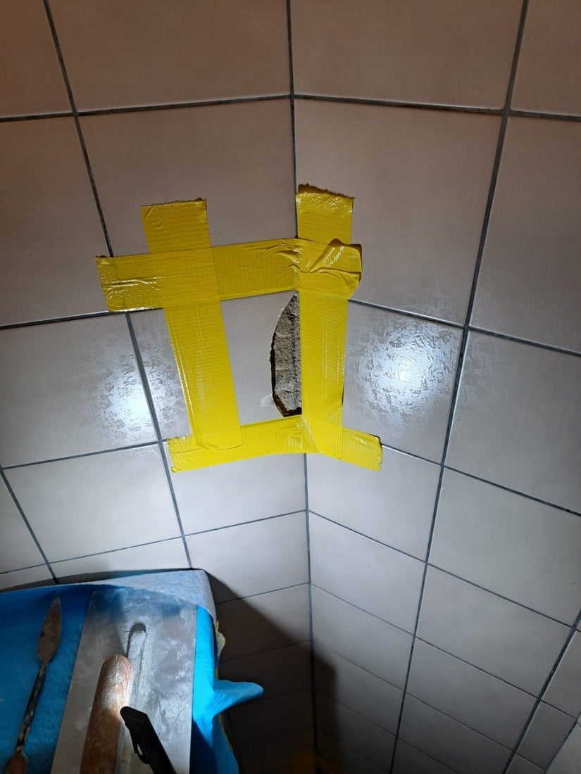 Defekte Wandfliese in Fliesenspiegel mit gelbem Klebeband provisorisch repariert, daneben Bauwerkzeuge.