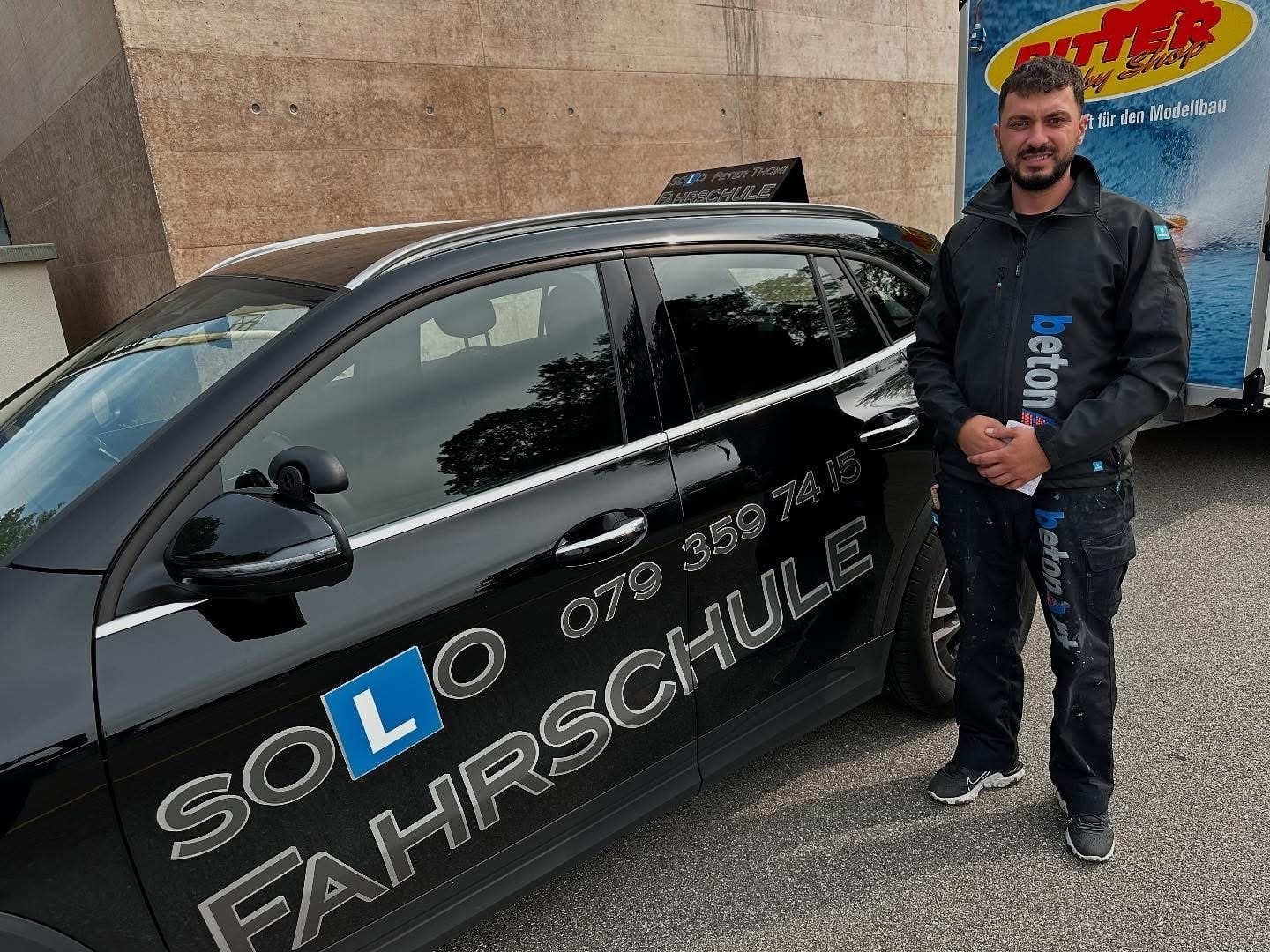 Mann steht neben einem schwarzen Auto mit Solo Fahrschule-Aufschrift auf dem Parkplatz, L-Schild deutlich sichtbar.