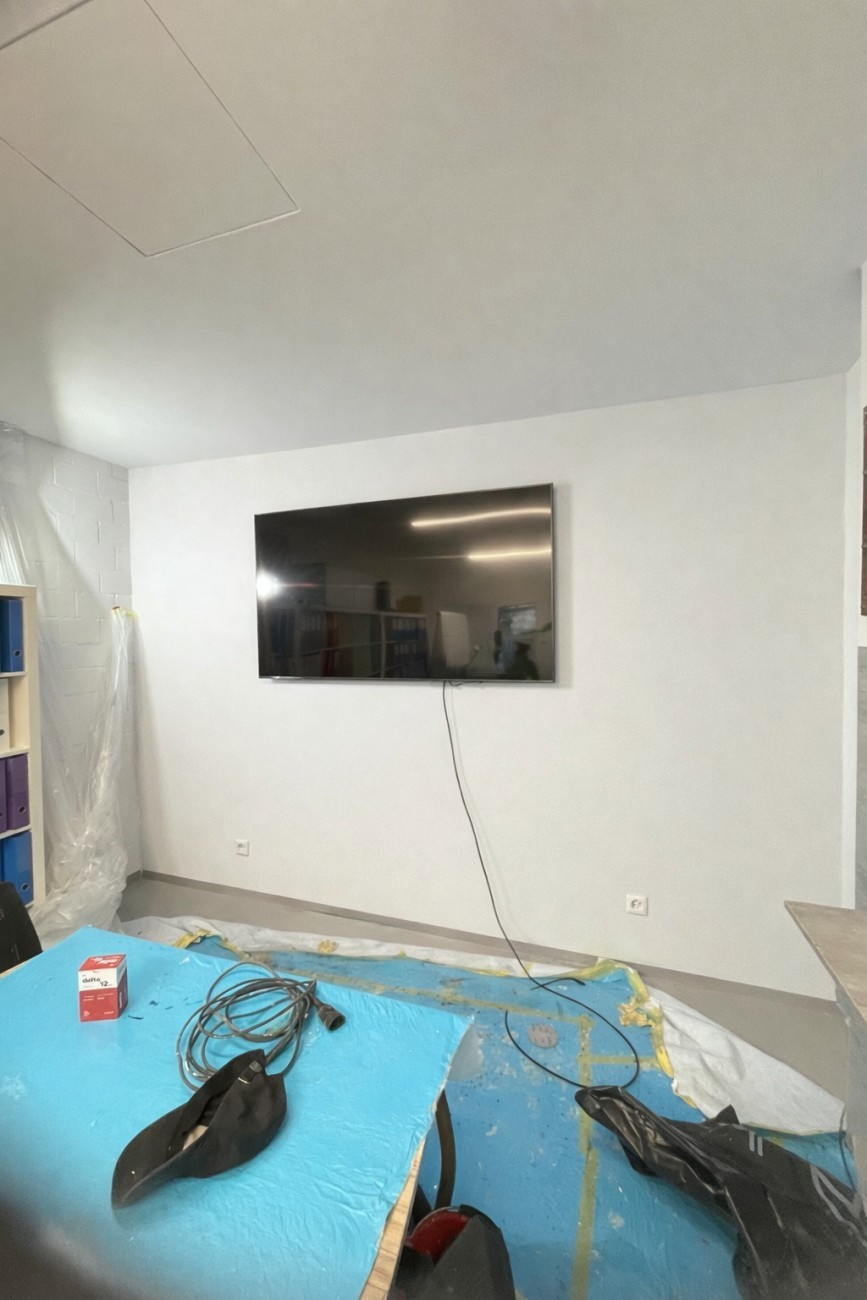 Großer Fernseher an weißer Wand in modernem Büro, Teppich und Werkzeuge auf blauem Abdeckvlies, Renovierung im Gange.